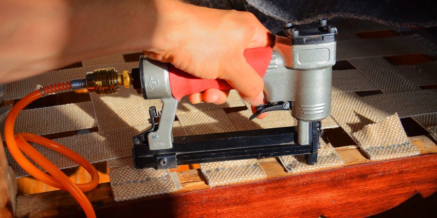 Hand holding a pneumatic staple gun over woven fabric on a wooden chair frame