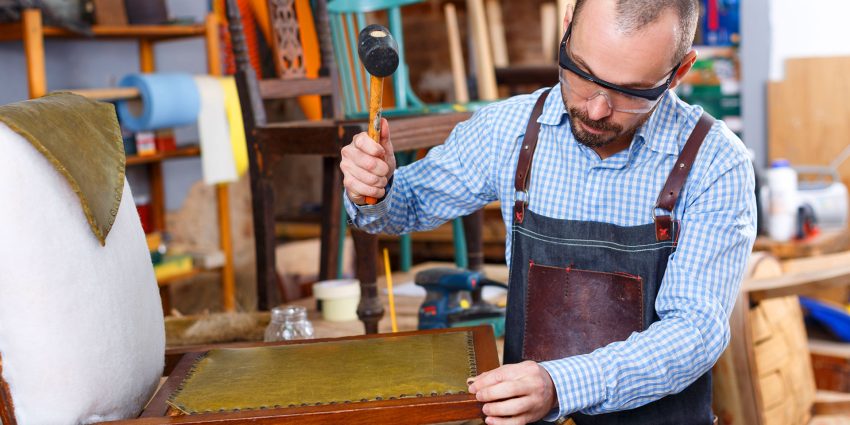 Upholsterer using a mallet to secure fabric onto a wooden chair frame in workshop